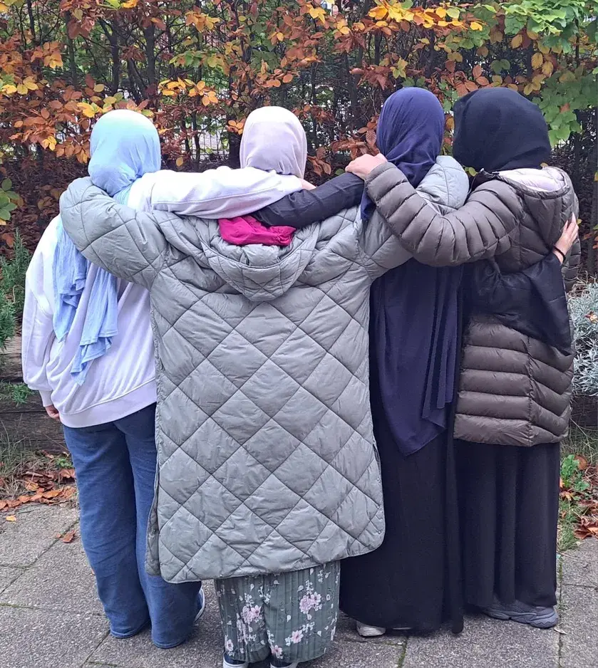 Four girls in headscarves standing arm in arm, seen from behind, in an autumn park