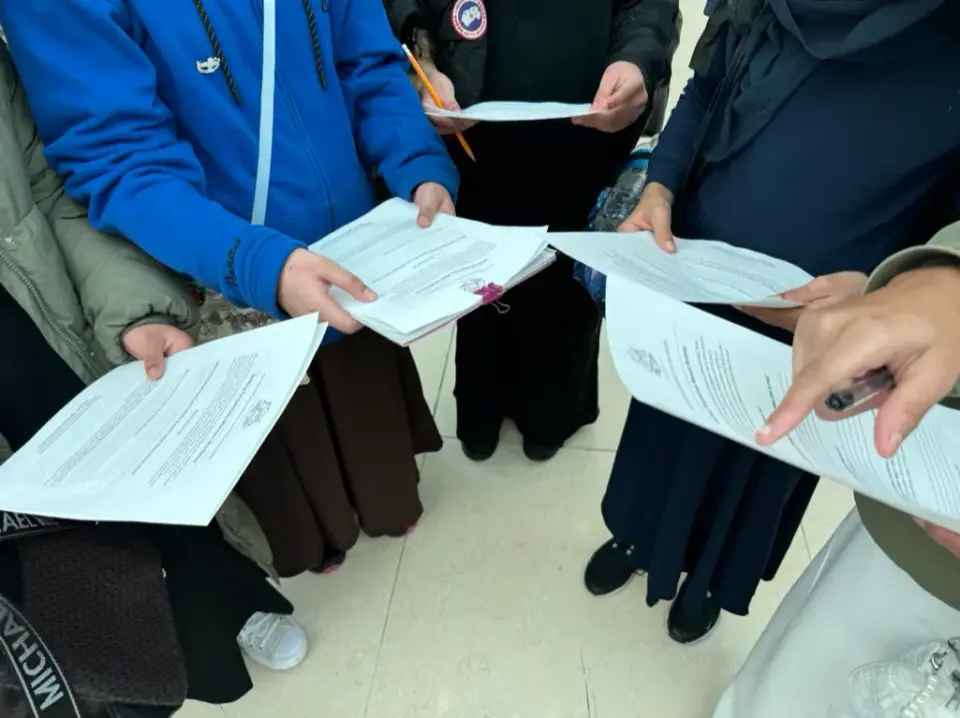 Students in headscarves standing in a circle, reading printed worksheets together