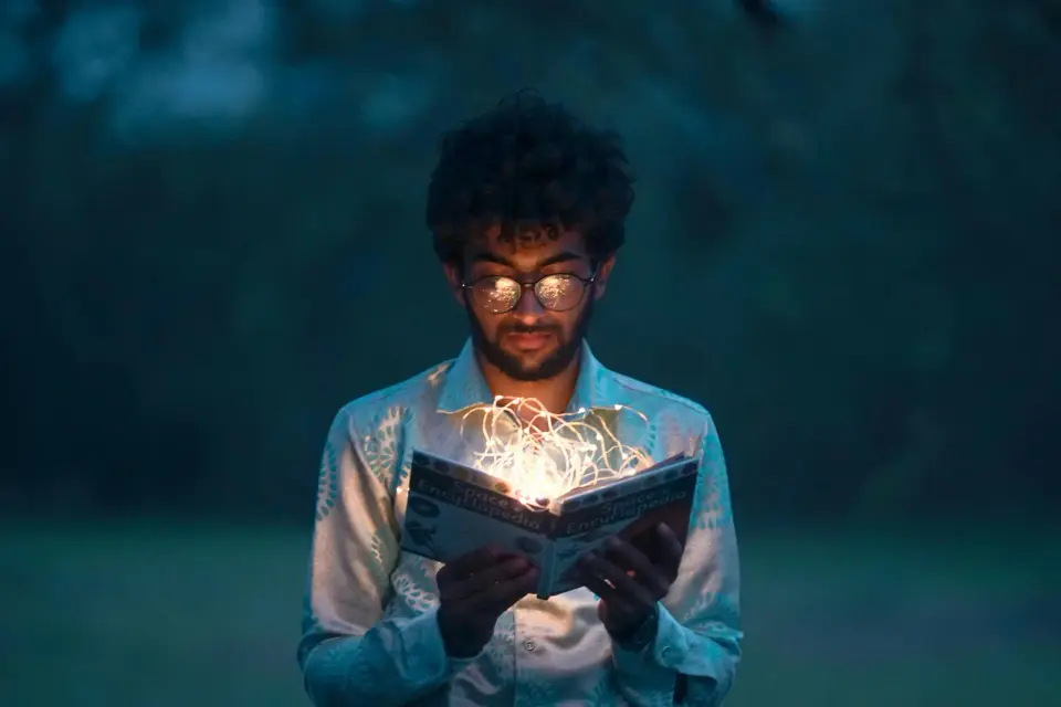 A young man reading a glowing book outdoors at dusk