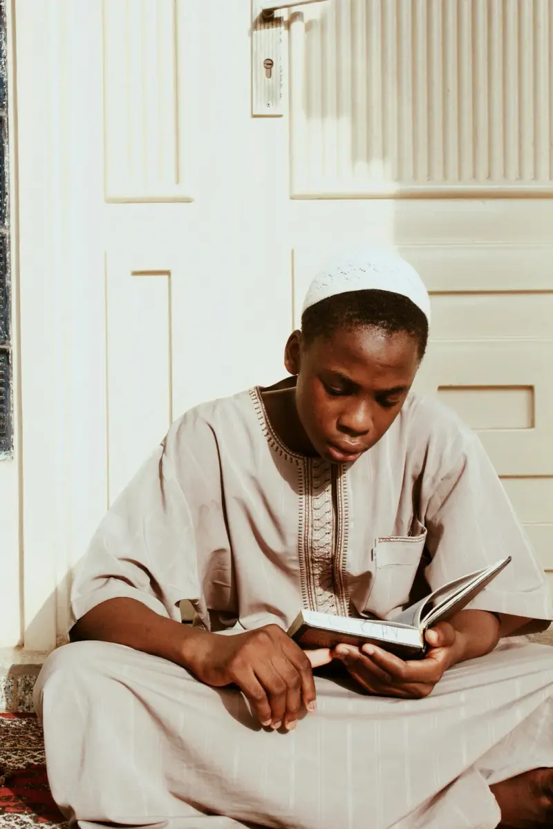 A boy in a white thobe and kufi reading the Qur'an