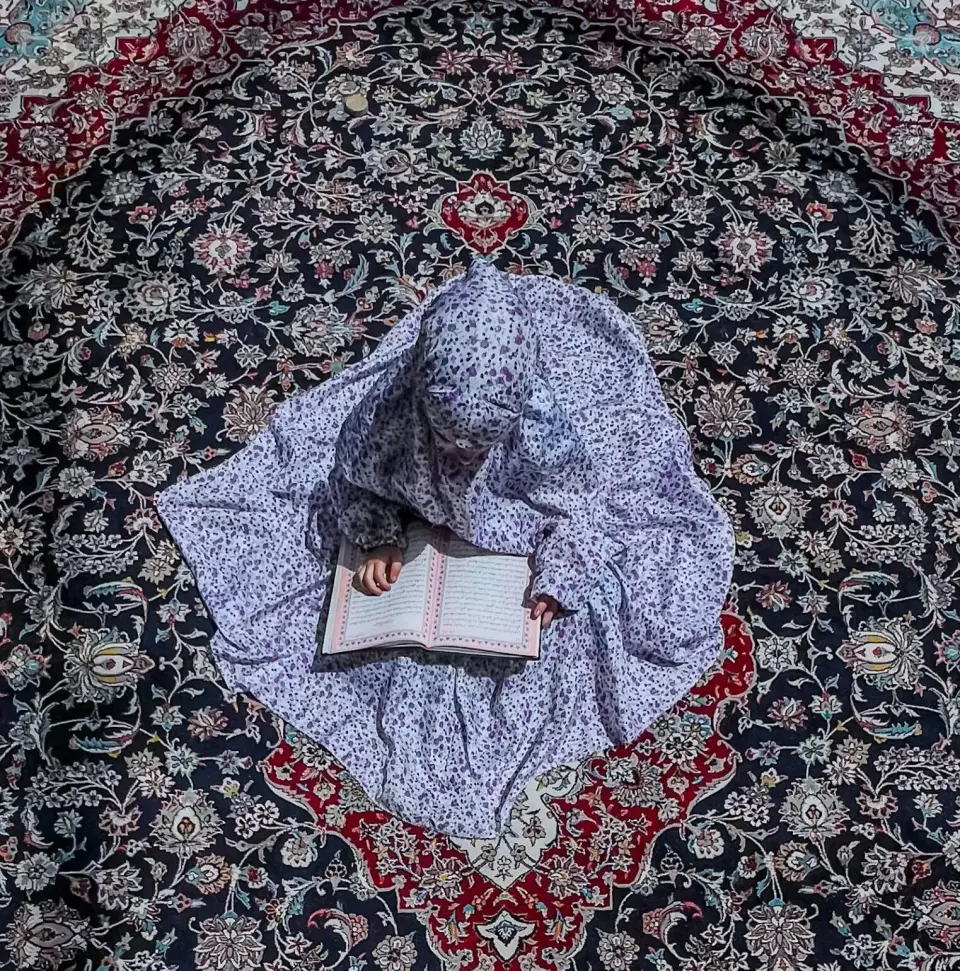 A girl in a prayer garment reading the Qur'an on a patterned rug, seen from above