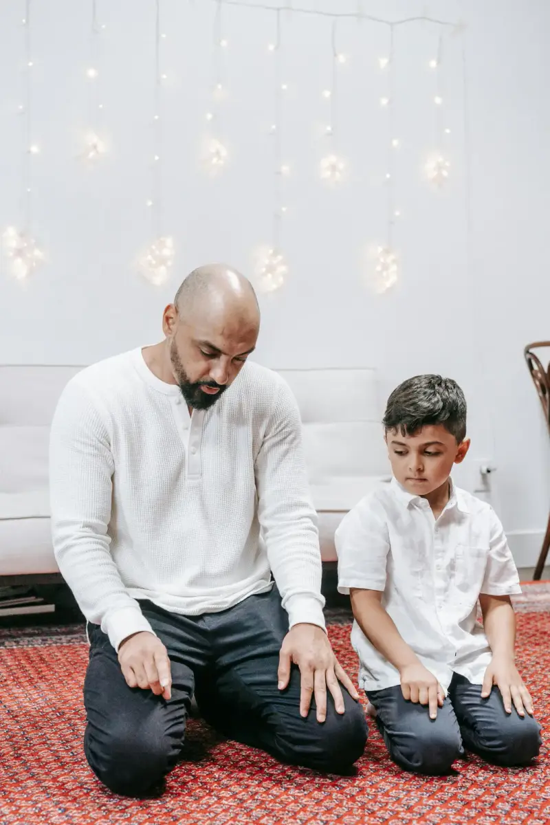 A father and son praying side by side on a prayer mat at home
